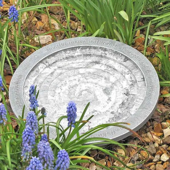 An artisanal birdbath on the ground with purple flowers in the foreground.