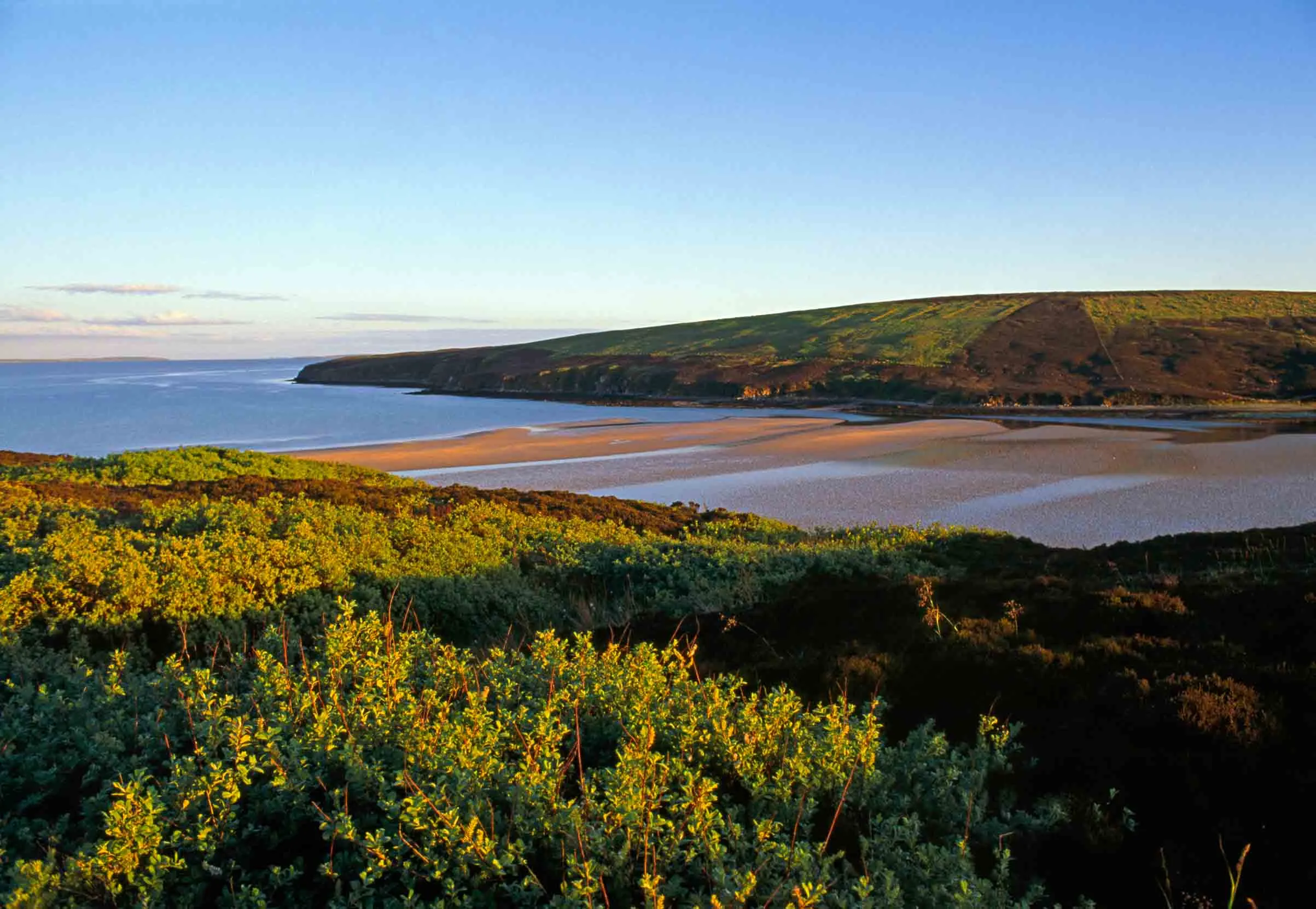 Sun sets over the bay reflecting the pink sky on the water as it flows onto the sandy beach, surrounded by grassy hillsides.