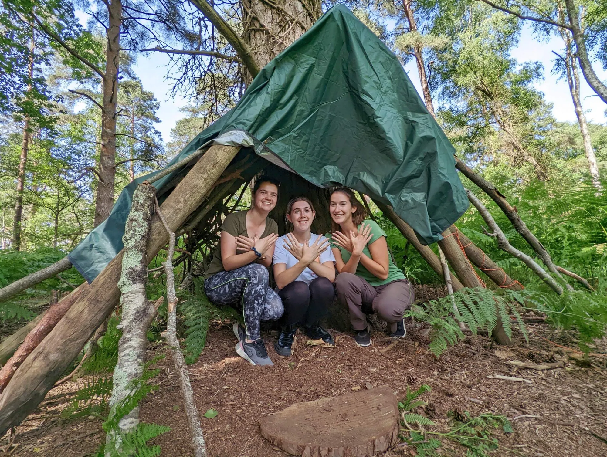 Three young people crouched in a shelter made from logs and tarp.