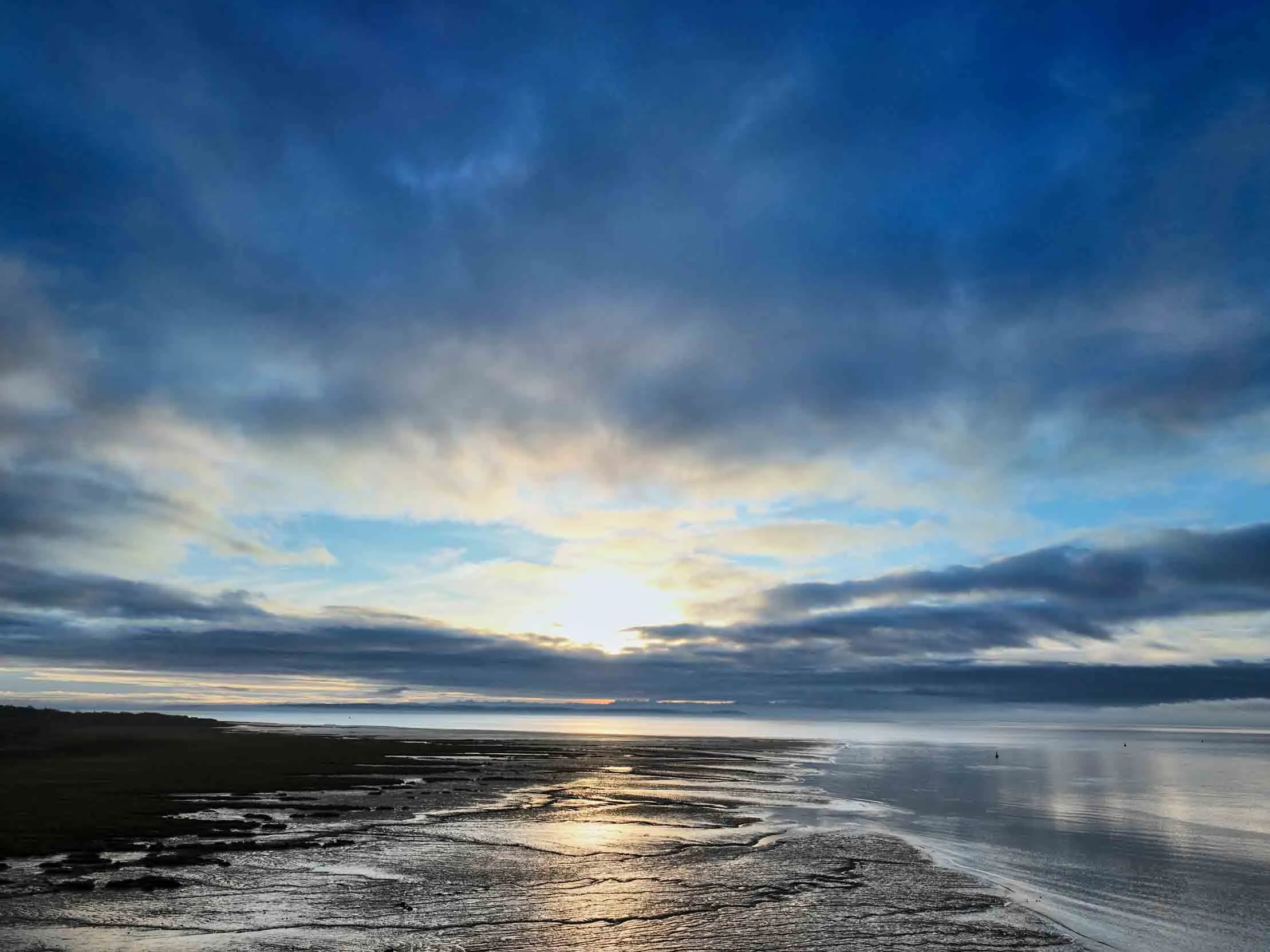 A moody blue sky with fluffy white clouds appears of the edge of a body of water breaking on the land.