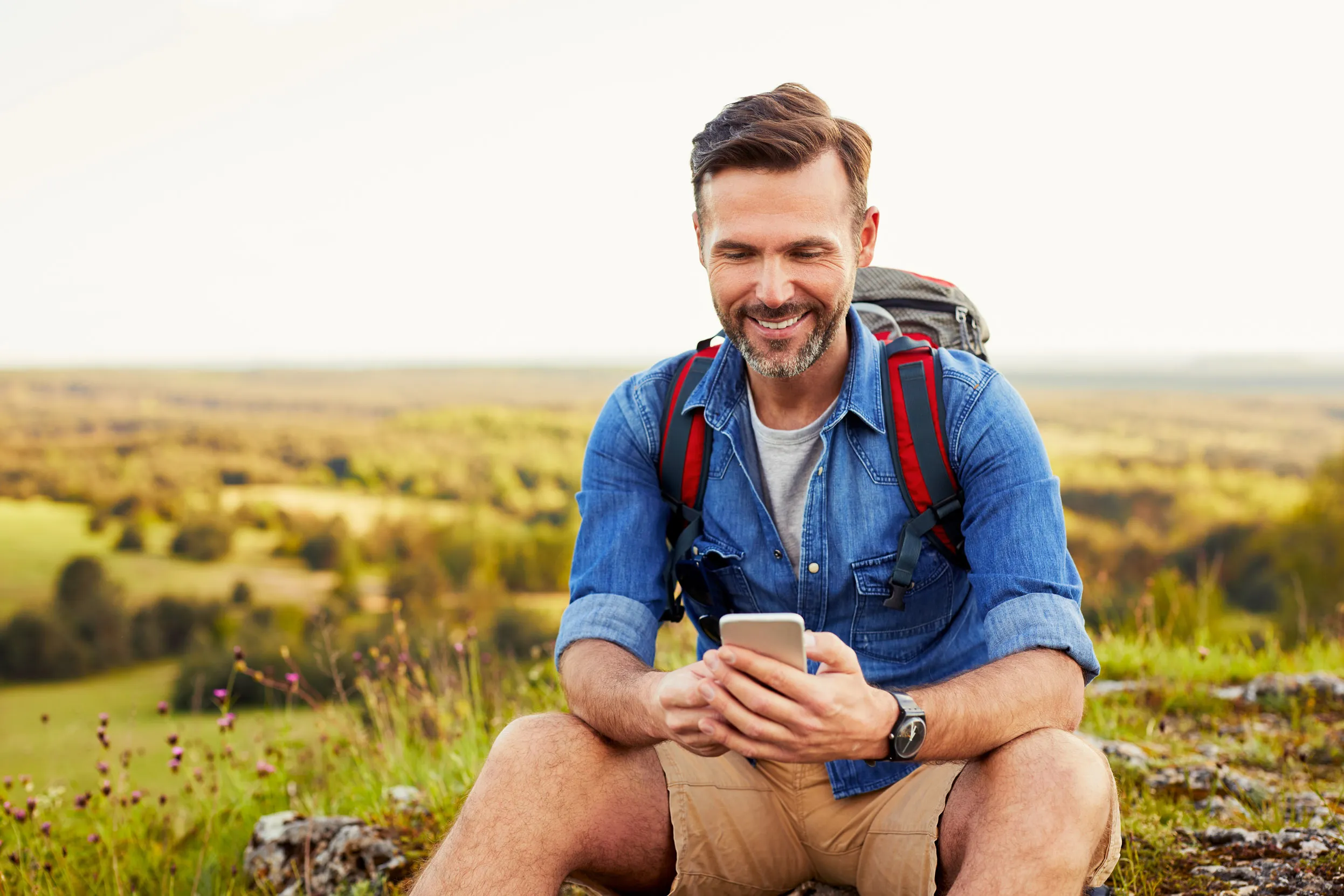 A person with a rucksack on using their phone, sat on a rock in front of a grassy landscape.