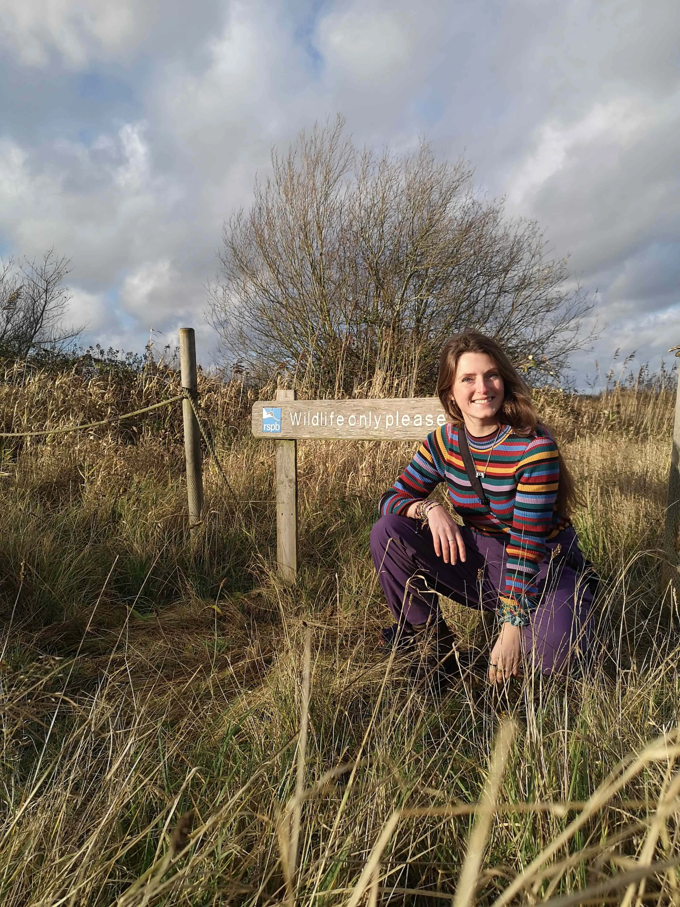 Lucy Hodson kneeling outdoors by a sign that reads "Wildlife Only Please" under a cloudy sky. 