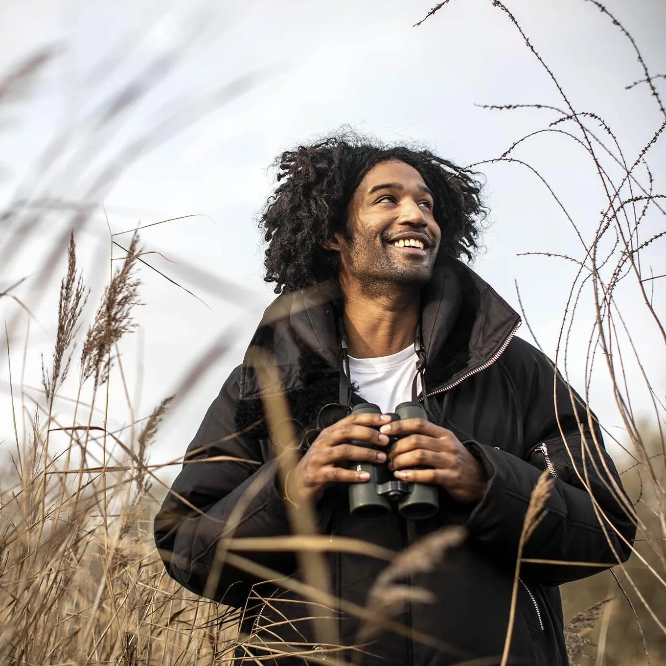 Nadeem Perera smiling while holding binoculars in a tall grassy field, enjoying the outdoors.