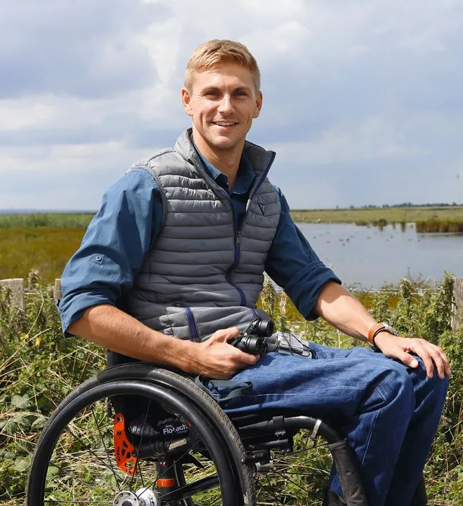 Steve Brown smiling while seated in a wheelchair, holding binoculars against a scenic wetland backdrop.